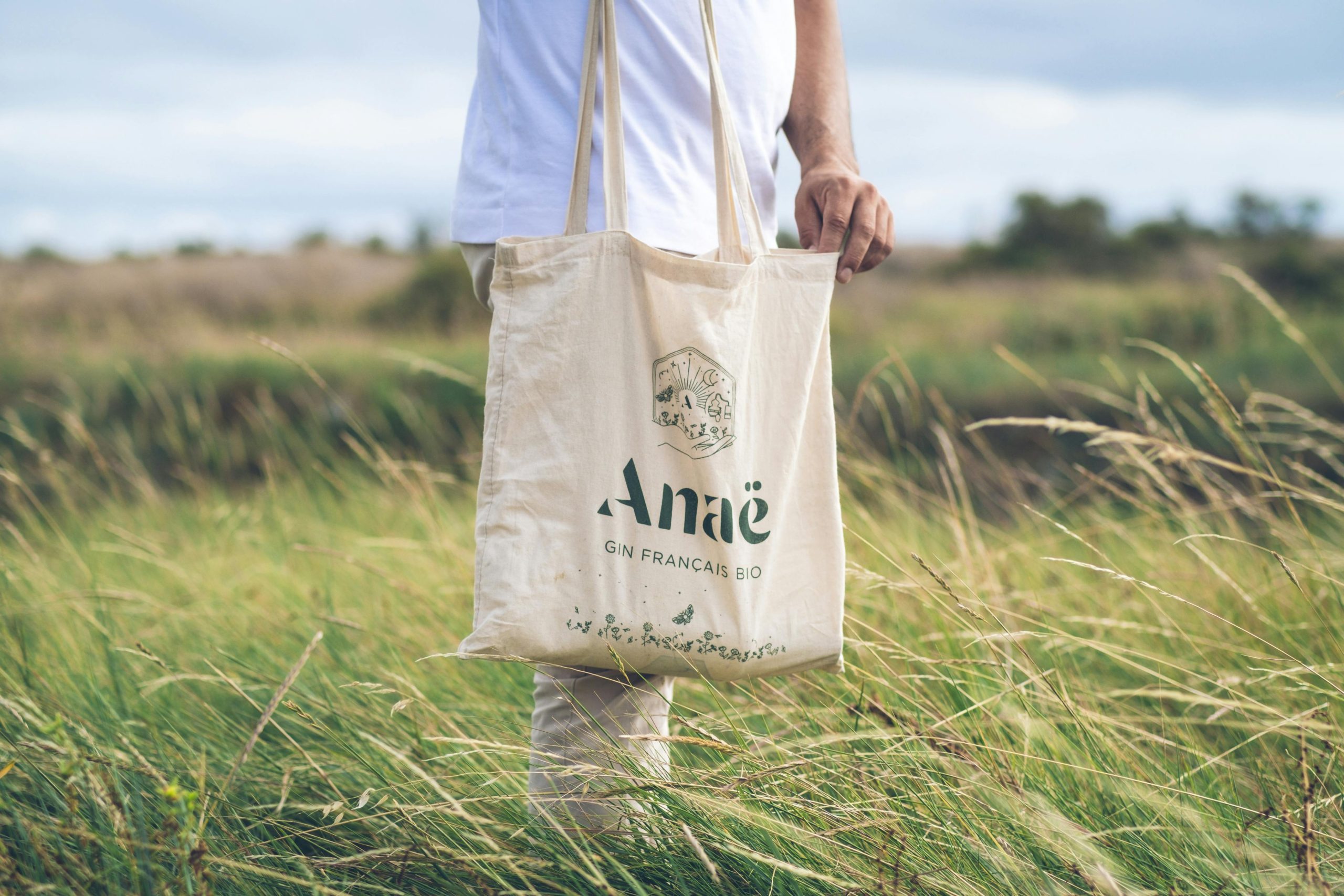 A person holding a canvas tote bag with Anaë logo in a green field, promoting sustainability.